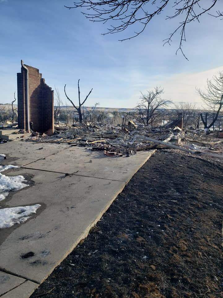 Picture of a burned structure surrounded by scorched trees. Toward the left of the photo is a brick wall standing on its own while burnt debris covers the lot.