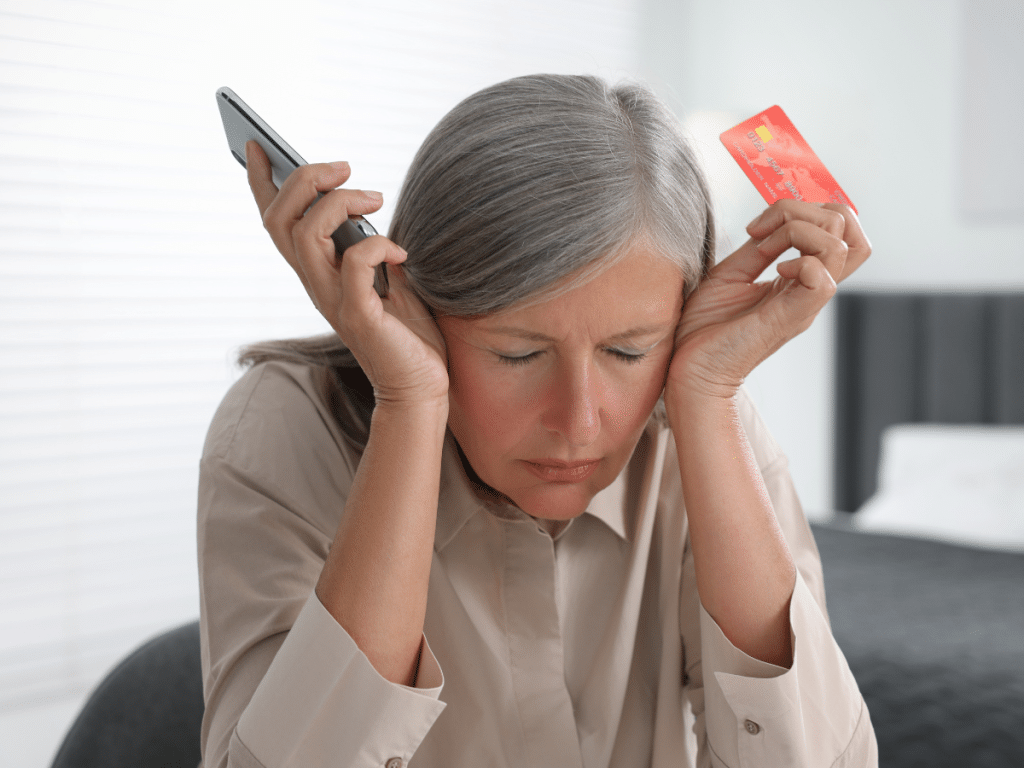 A lady with gray hair holds her phone and credit card looking frustrated by an online air duct cleaning scam. 